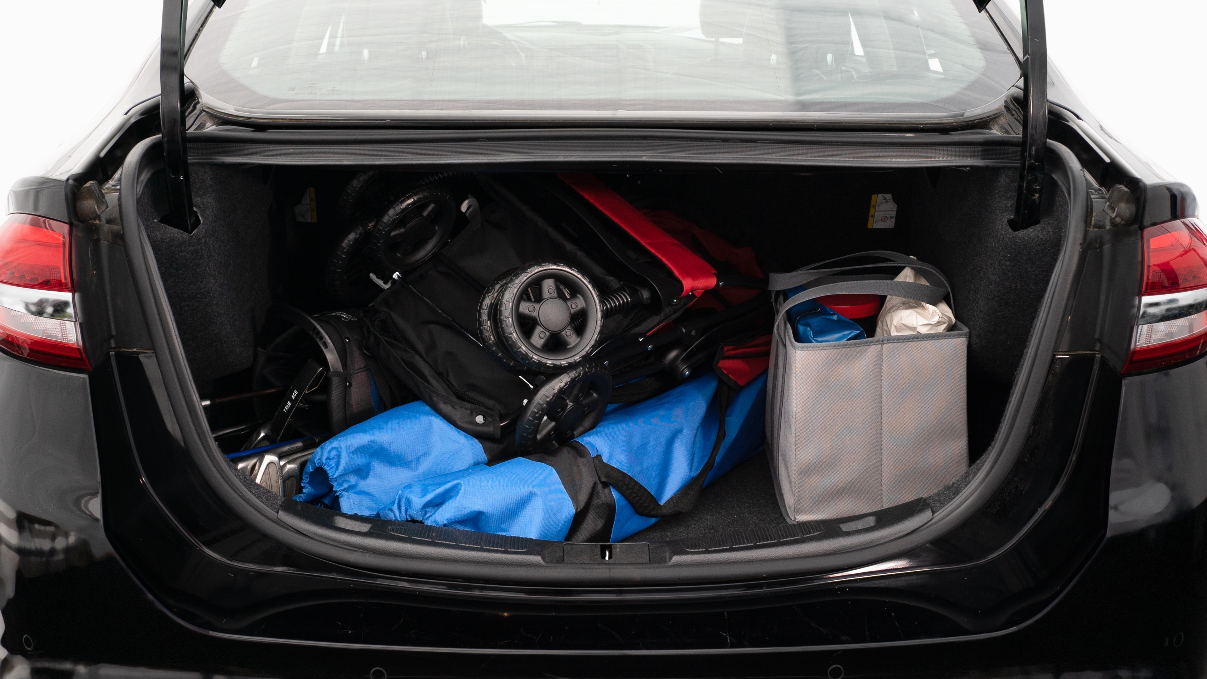 The trunk lid is open showing full view of the trunk. A baby stroller, two grocery carriers, two camping chairs in the carrying bag, and a set of golf clubs are placed inside.