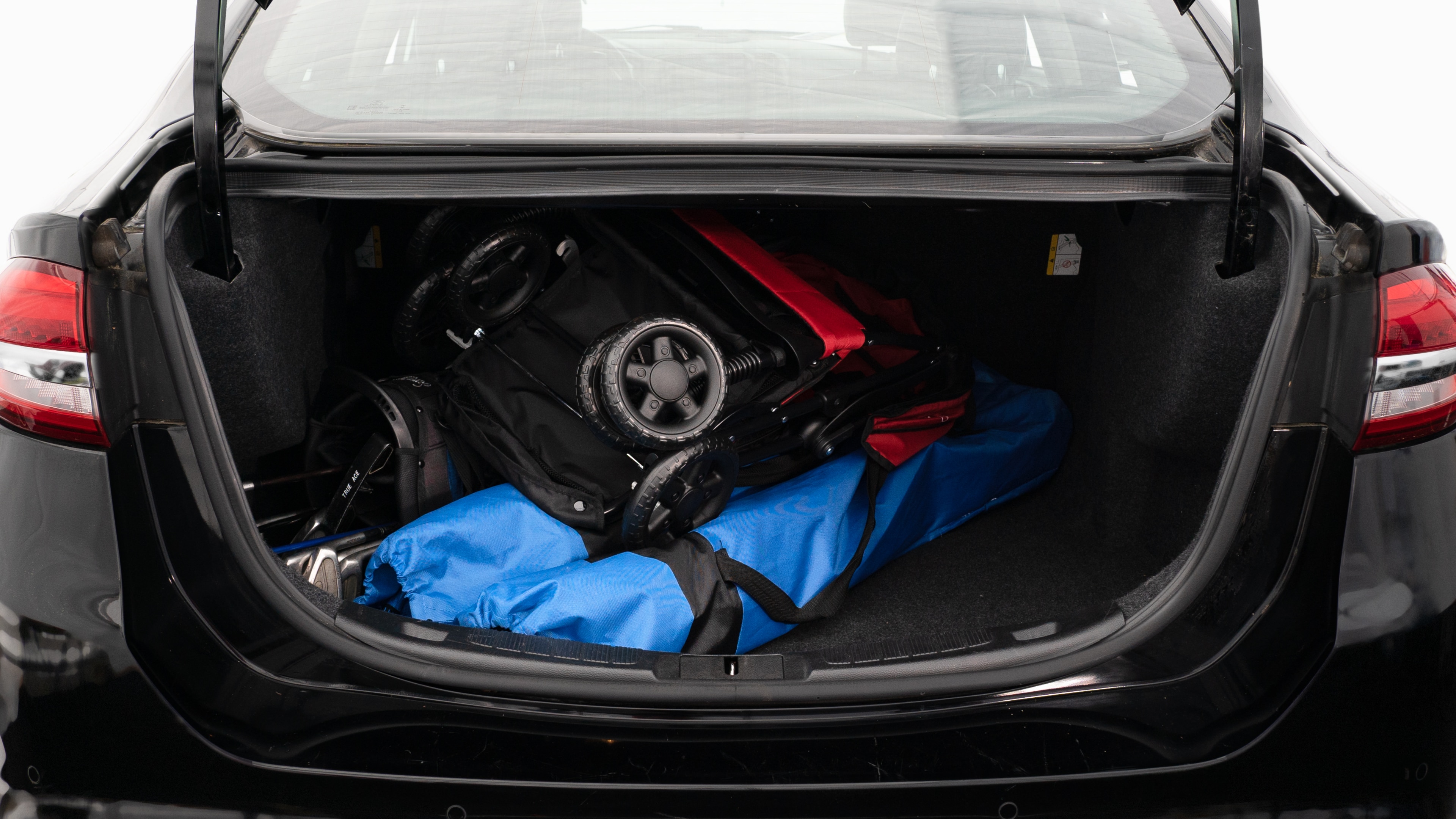 The trunk lid is open showing full view of the trunk. A baby stroller, two grocery carriers, two camping chairs in the carrying bag, and a set of golf clubs are placed inside.
